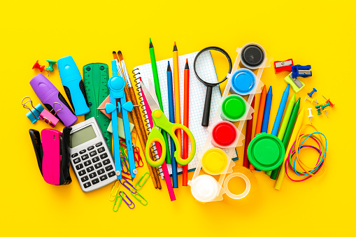 Large group of multi colored school supplies shot on yellow background. The composition includes a drawing compass, color pencils, paint brushes, paper clips, pencil sharpener, eraser, crayons, pocket calculator, thumbtack, rubber bands, notepad, stapler, adhesive tape, felt tip pen and scissors.