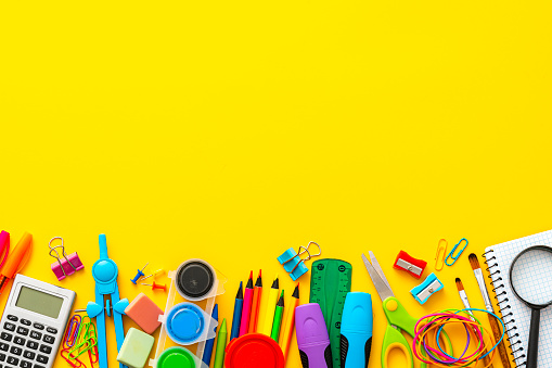 Large group of multi colored school supplies arranged at the bottom of a yellow background. Copy space. The composition includes a drawing compass, color pencils, paint brushes, paper clips, pencil sharpener, eraser, crayons, pocket calculator, thumbtack, rubber bands, notepad, stapler, adhesive tape, felt tip pen and scissors.
