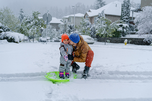 Portrait of a young father squatting down next to his multiracial two year old daughter and affectionately giving her a kiss on the cheek while playing together in the snow in their residential neighborhood. Both individuals are bundled up in winter clothing and smiling directly at the camera.