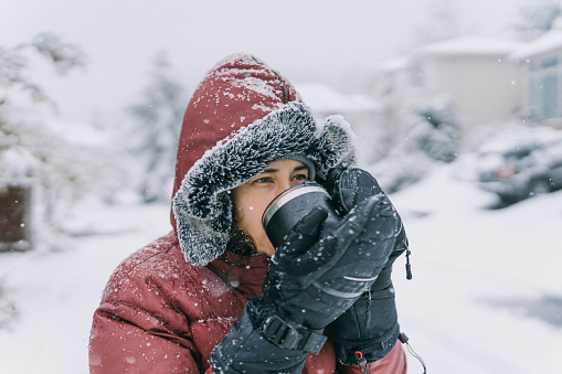 Young multiracial woman of Hawaiian and Chinese descent is bundled up in winter clothing, drinking a hot beverage out of a travel mug, while standing outside in her neighborhood during a snowstorm.