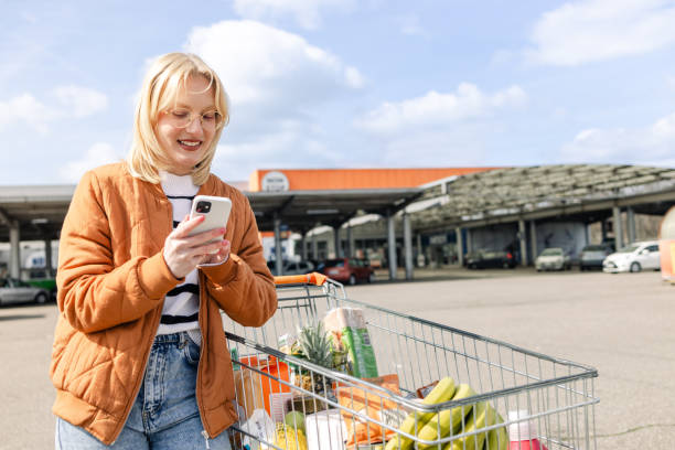istock Young woman using her phone, while standing next to a shopping cart 1574599850