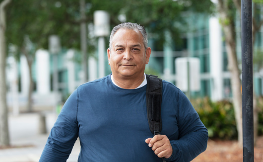 A mature Hispanic man, in his 50s, walking in the city carrying a backpack. He is looking at the camera with a serious expression.