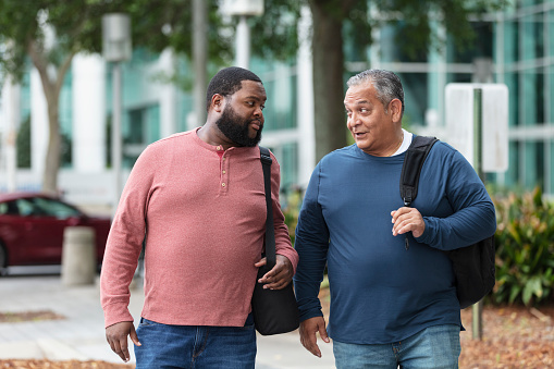Two heavyset multiracial men walking side by side, conversing, in the city outside a building, carrying backpacks. The man with the beard is African-American, in his 30s. His friend is Hispanic, in his 50s.