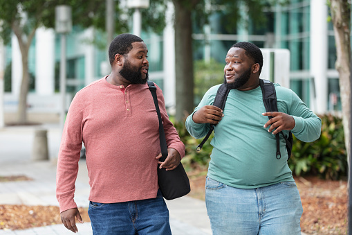 Two plus size bearded men walking side by side on a city sidewalk, outside a building, conversing. The one on the right carrying a backpack is an young man in his 20s, mixed race, black and Pacific Islander. His African-American friend is in his 30s.