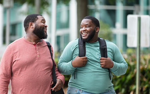 Two plus size bearded men walking side by side on a city sidewalk, outside a building, conversing. The one on the right carrying a backpack is an young man in his 20s, mixed race, black and Pacific Islander. His African-American friend is in his 30s.