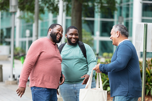 A multiracial group of three plus size men conversing and laughing on a sidewalk outside a building. The mature man on the right is Hispanic, in his 50s. The young man in the middle is mixed race, black and Pacific Islander, in his 20s. Their friend is African-American, in his 30s.
