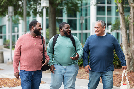 A multiracial group of three plus size men walking on a sidewalk outside a building, smiling and conversing. The mature man on the right is Hispanic, in his 50s. The young man in the middle is mixed race, black and Pacific Islander, in his 20s. Their friend is African-American, in his 30s. They are all carrying bags.