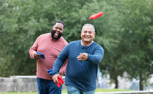 Two plus size multiracial men having fun at the park playing a bean bag toss game. A mature Hispanic man, in his 50s, has just thrown a bean bag. The main focus is on his African-American friend, in his 30s, standing behind him, shouting.