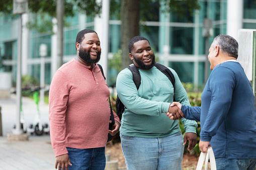 A multiracial group of three plus size men meeting and greeting on a sidewalk outside a building, smiling and shaking hands. The mature Hispanic man on the right is out of focus. He is shaking hands with a young black and Pacific Islander man, while their African-American friend watches.