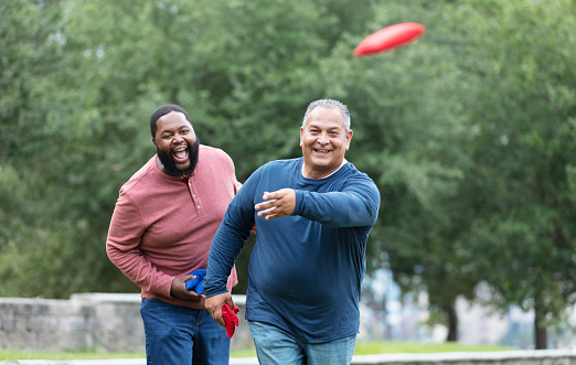 Two plus size multiracial men having fun at the park playing a bean bag toss game. The main focus is on the mature Hispanic man, in his 50s, who has just thrown a bean bag. His African-American friend, in his 30s, is standing behind him, shouting, perhaps trying to distract him.