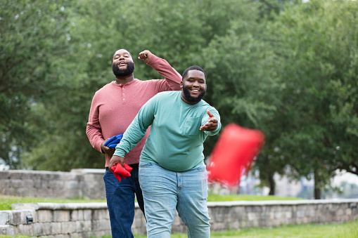 Two plus size men having fun at the park playing a bean bag toss game. The main focus is on the young man throwing the bean bag. He is mixed race, black and Pacific Islander, in his 20s. His African-American friend is behind him, watching and punching the air. cheering a good shot.