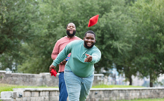 Two plus size men having fun at the park playing a bean bag toss game. The main focus is on the young man throwing the bean bag. He is mixed race, black and Pacific Islander, in his 20s. His African-American friend is behind him, watching, waiting his turn.