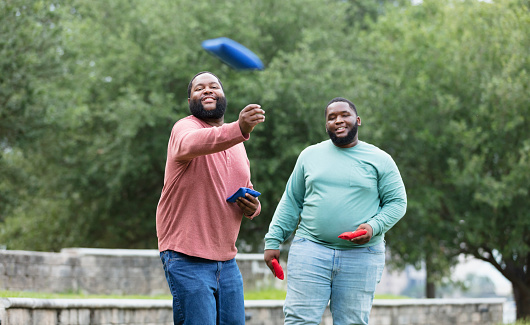 Two plus size men having fun at the park playing a bean bag toss game. The main focus is on the man throwing the bean bag. He is African-American, in his 30s. His friend is behind him, watching, waiting his turn.