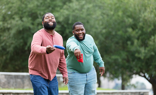 Two plus size men having fun at the park playing a bean bag toss game. The main focus is on the man throwing the bean bag. He is African-American, in his 30s. His friend is behind him, laughing, trying to distract him.