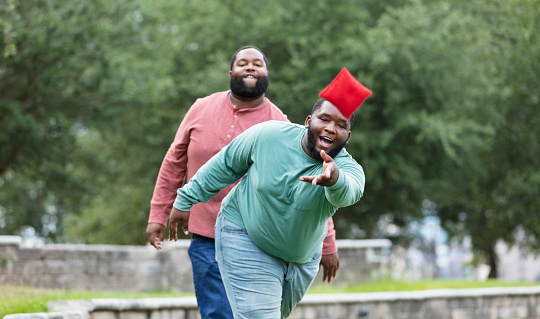 Two plus size men having fun at the park playing a bean bag toss game. The main focus is on the young man throwing the bean bag. He is mixed race, black and Pacific Islander, in his 20s. His African-American friend is behind him, watching, making a face.