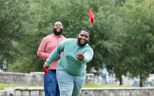 Two plus size men having fun at the park playing a bean bag toss game. The main focus is on the young man throwing the bean bag. He is mixed race, black and Pacific Islander, in his 20s. His African-American friend is behind him, watching, waiting his turn.