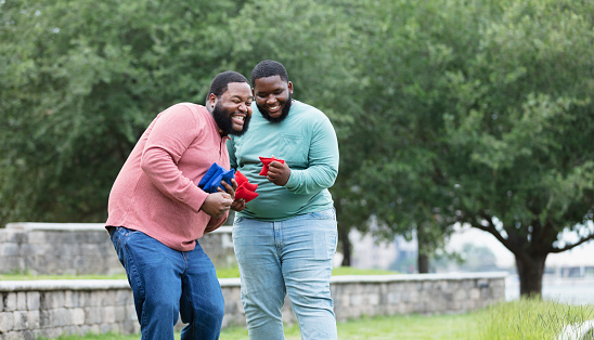 Two plus size men having fun at the park playing a bean bag toss game. The main focus is on the young man looking down at the bean bag in his hand. He is mixed race, black and Pacific Islander, in his 20s. His African-American friend is winking, making a fist, and either grinning or gritting his teeth, so it's hard to tell if his toss was good.