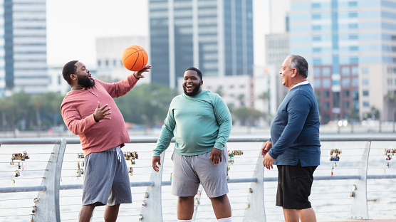 A multiracial group of three plus size men have fun playing basketball on a city waterfront. The African-American man on the left, in his 30s, is showing off his ball-handling skills.