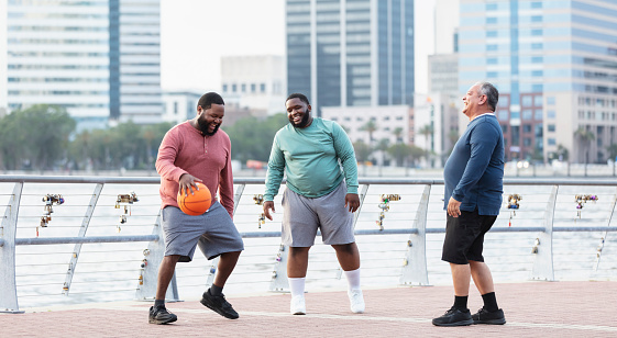 A multiracial group of three plus size men have fun playing basketball on a city waterfront. The African-American man on the left, in his 30s, is dribbling the ball while his friends laugh.