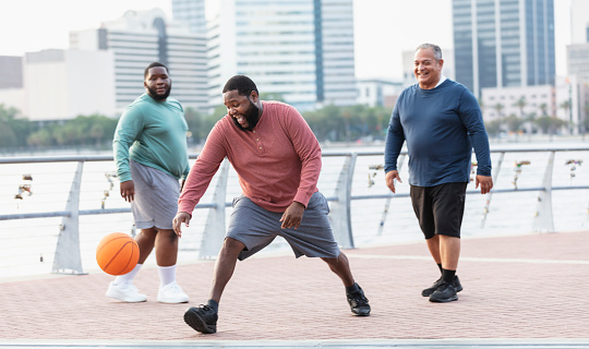 A multiracial group of three plus size men have fun playing basketball on a city waterfront. The focus is on the African-American man reaching for the ball. He is in his 30s.