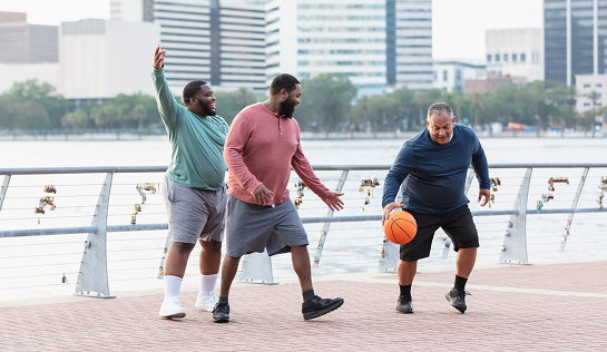 A multiracial group of three plus size men have fun playing basketball on a city waterfront. The man dribbling the ball is Hispanic, in his 50s. The two black men are in their 20s and 30s.