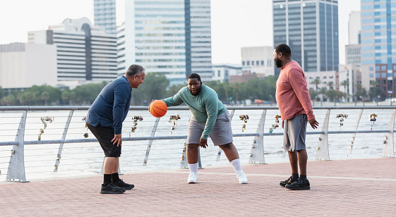 A multiracial group of three plus size men have fun playing basketball on a city waterfront. The man dribbling the ball is mixed race, black and Pacific Islander, in his 20s. He is looking toward the camera. The African-American man on the right is in his 30s, and their Hispanic friend is in his 50s.