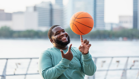 A young man standing by a city waterfront balancing a basketball on his fingertip. He is multiracial, black and Pacific Islander, in his 20s.