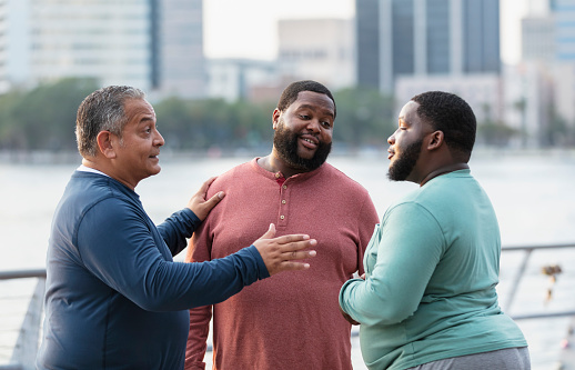 A multiracial group of three plus size men conversing on a city waterfront. The Hispanic man on the left is in his 50s. The African-American man in the middle is in his 30s. They are both looking at their younger friend, a mixed race, black and Pacific Islander, in his 20s. Perhaps they are offering him some advice.
