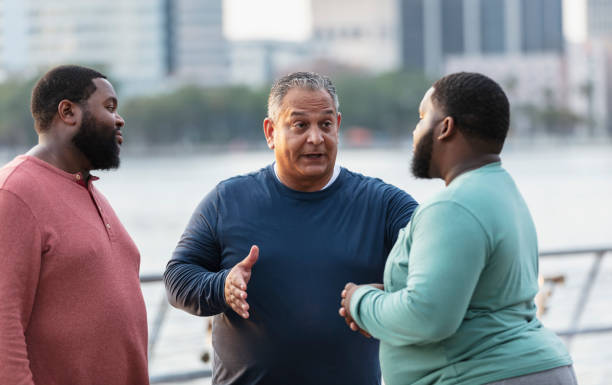 A multiracial group of three plus size men conversing on a city waterfront. The Hispanic man in the middle is in his 50s. He is speaking to the young man on the right, who is mixed race, black and Pacific Islander, in his 20s, perhaps offering him some advice. Their African-American friend on the left is in his 30s.
