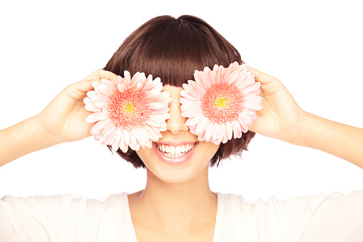 A happy young woman covering eyes with fresh pink Gerber Daisy flowers, isolated on white