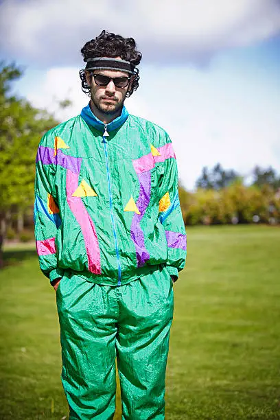 A man in a bright fluorescent sweat suit stands glumly with a sad, serious facial expression in a city park on a warm summer day.  He's decked out with a sweat band and tinted vintage glasses.