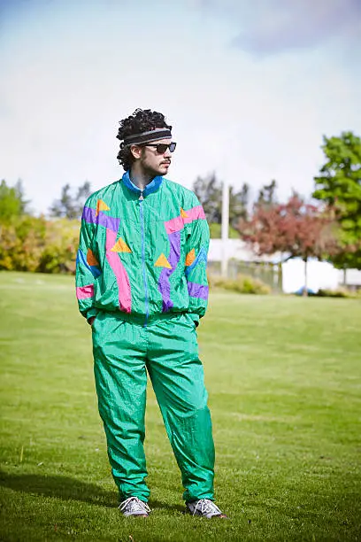 A man in a bright fluorescent sweat suit stands looking cool with a  serious facial expression in a city park on a warm summer day.  He's decked out with a sweat band and tinted vintage glasses.  Vertical with copy space.