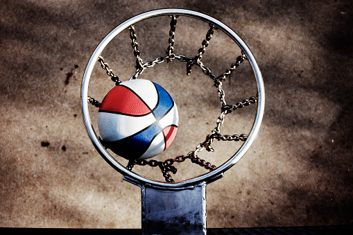 High contrast photo of a red, white and blue basketball dropping into a chain net in the city. 
