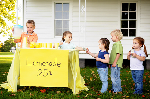 Color photo of a young boy and girl serving neighborhood kids at their lemonade stand.
