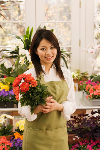 A small business owner of a retail flower shop. An Asian woman florist entrepreneur shopkeeper. She is working next to the checkout counter in her shop, posing and smiling at the camera, and surrounded by colorful garden center plants. Photographed in vertical format with copy space.
