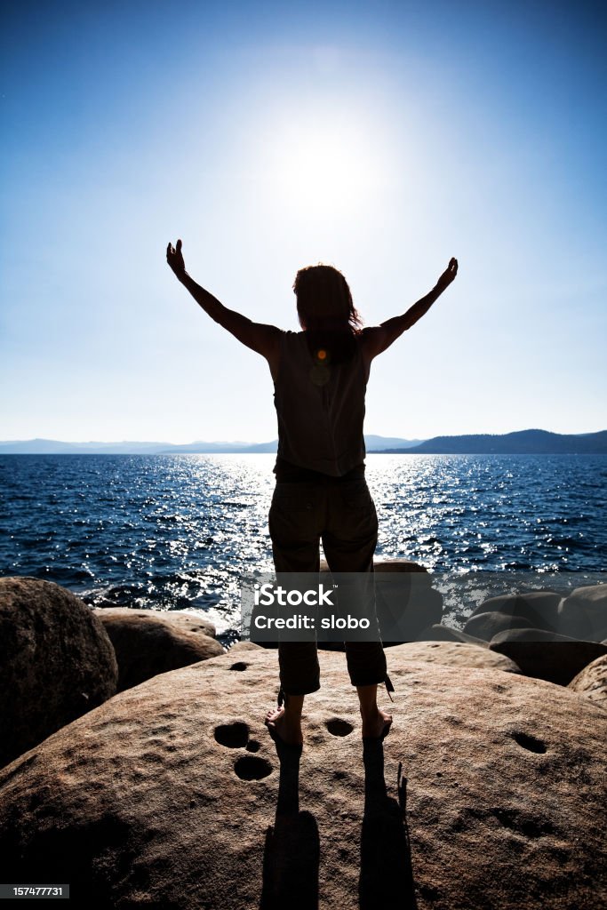 Praise the sun Woman standing on rocky shore with arms outstretched toward sun. Adult Stock Photo