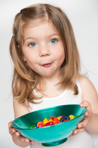 A cute little girl is looking straight at the camera trying to hide the candy in her mouth! She is holding a turquoise bowl of jellybeans. She is wearing a white sleeveless dress, and her mid-long hair is loose around her pretty face. Shallow DOP, focus on her blue eyes. Vertical with white background.