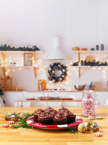 Chocolate brownie cupcakes frosted with chocolate frosting on a red plate on butcher block counter top.  Cupcakes have been sprinkled with peppermint candy.  Kitchen is decorated for Christmas and ornaments are on the counter beside the plate of cupcakes.