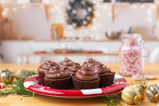 Chocolate brownie cupcakes frosted with chocolate frosting on a red plate on butcher block counter top.  Cupcakes have been sprinkled with peppermint candy.  Kitchen is decorated for Christmas and ornaments are on the counter beside the plate of cupcakes.