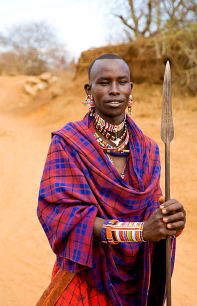 Young Masai warrior with spear and traditional dress, Kenya. Young Masai warrior (morani) with spear. Dressed in traditional checkered red shuka and pearl jewelry and carrying a spear with the blade upwards.  It is dry season and the red soil is very dry, no leaves on the trees in the background. Vertical composition. The area is the Selenkay Conservancy close to Amboseli National Park in southern Kenya, East Africa kenyan man stock pictures, royalty-free photos & images