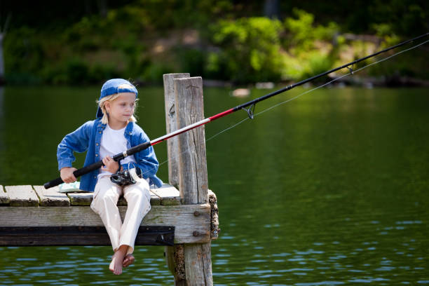 A 7-year-old girl sits with the fishing rod on the jetty at a lake and waits for a fish to bite. She looks into the camera and splashes her feet in the water