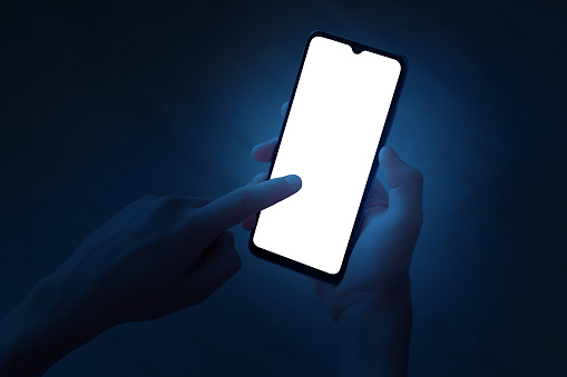 A teenager holds a smartphone with one hand, and touches the device's screen with his index finger at night. Phone with white screen on defocused dark blue background. There is a work path in the file.