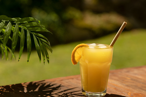 A glass of fresh orange juice on a wooden table under a palm tree branch outside in summer