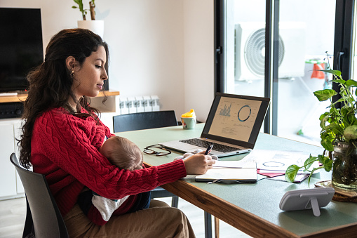 Young mom working home office on laptop while hold her baby on lap at home