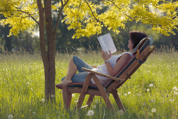 istock Young woman on wooden deck chair under tree reading book 157482400