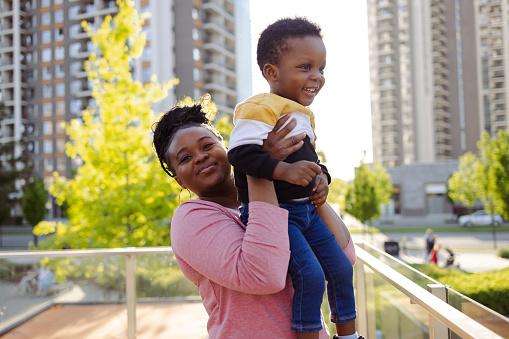 A young African American woman is looking at the camera, while cheerfully lifting up her young son in the air.