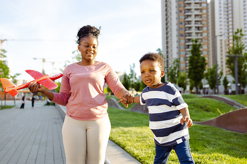 A young African American woman is holding her young son's hand, while out on a walk. She's holding his plane toy.