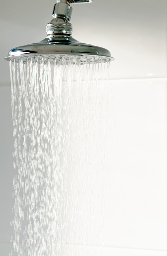 Water falling from a modern, chrome shower head against a background of white bathroom tiles.