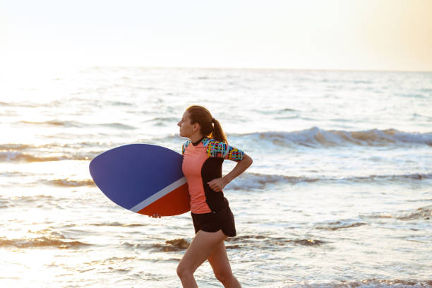 istock Young Caucasian woman running on the beach, while holding a skimboard 1574928759