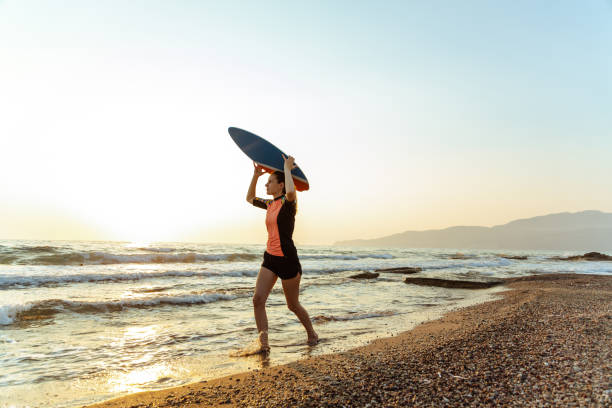 頭の上にスキムボードを持ちながらビーチを走る若い白人女性 - skimboard ストックフォトと画像